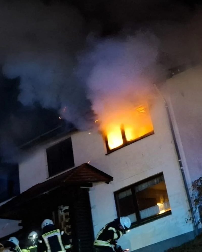 Nächtlicher Wohnhausbrand: Flammen schlagen aus einem Fenster im Obergeschoss, Feuerwehrleute arbeiten am Hauseingang